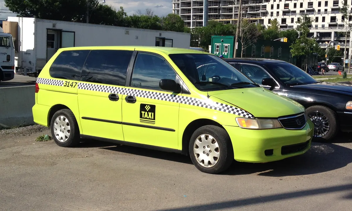 Lime-green minivan taxi with a black-and-white checkered stripe and a taxi sign on the door, parked in a lot